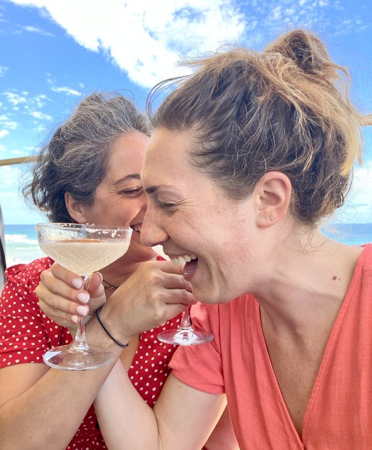 Two women laughing and sharing a drink with ocean view at Curl Curl Beach