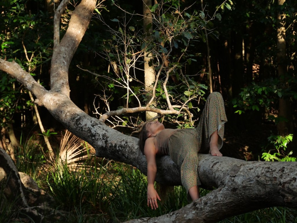 Woman standing in forest with arms open, surrounded by trees and greenery